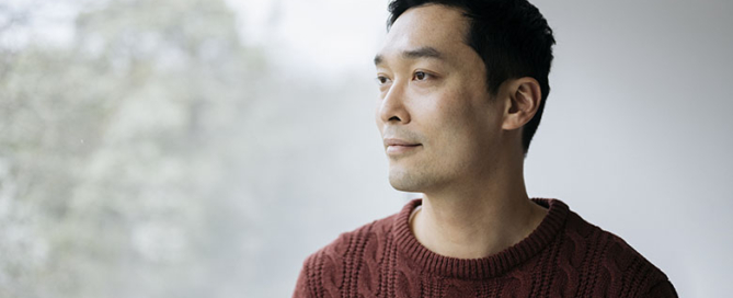 Man with short black hair in rust colored cable stitch sweater standing at picture window and looking away from camera with contented expression.