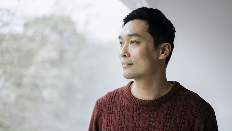 Man with short black hair in rust colored cable stitch sweater standing at picture window and looking away from camera with contented expression.